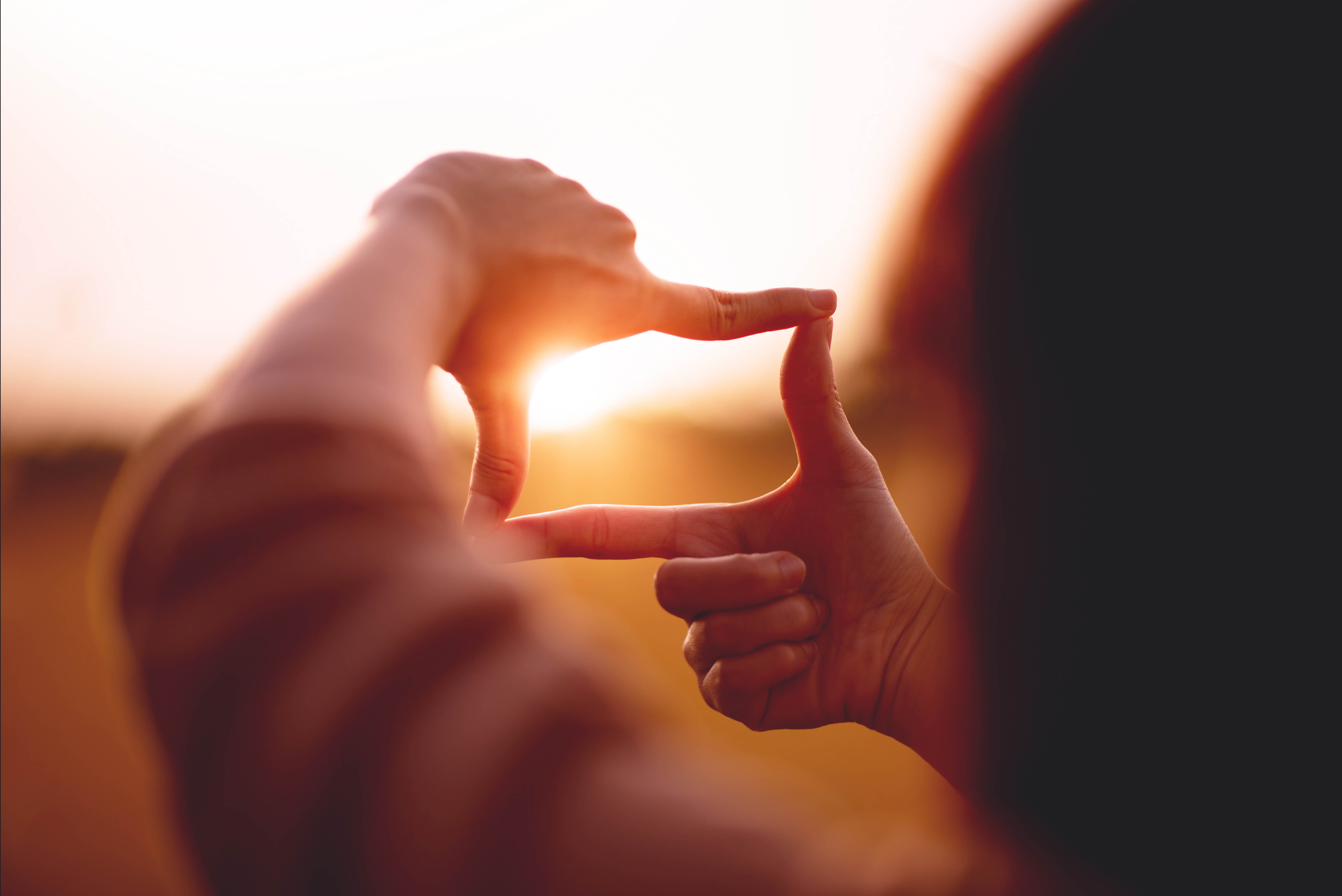 Woman holding up her hands in the shape of a rectangle, framing the sunset in the middle like she's preparing to take a photo